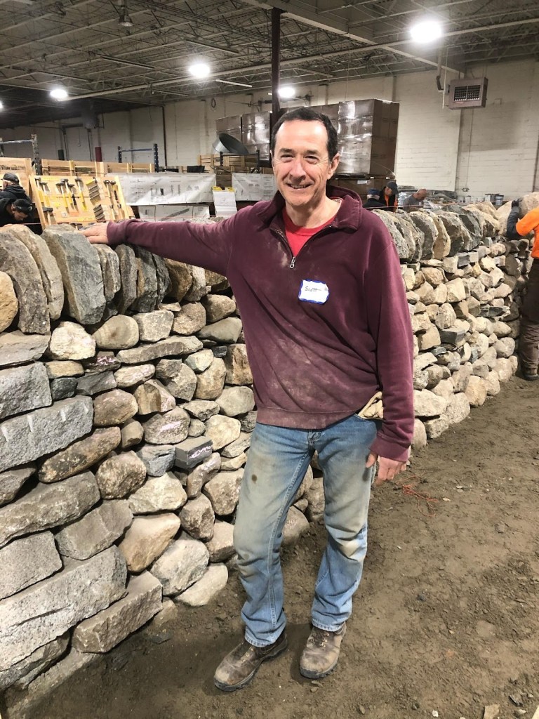 A man standing next to a stone wall in a workshop, smiling and wearing a maroon shirt with a name tag. The background shows tools and other stonework projects.