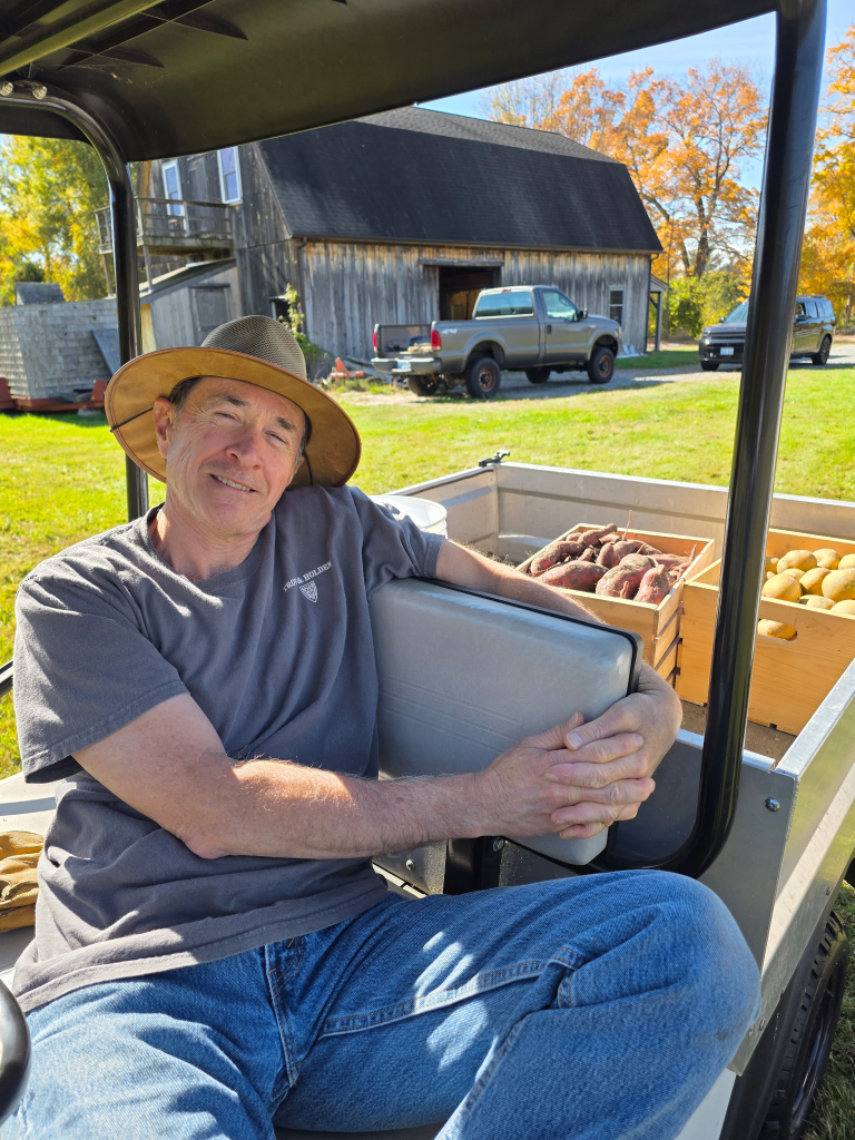 A man sitting on a cart with sweet potatoes and potatoes in wooden crates, set against a backdrop of trees and a rustic barn.