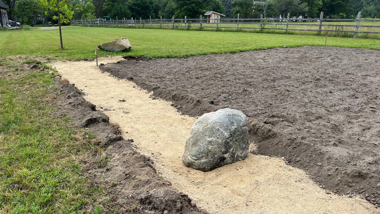 A partially constructed pathway made of sand between grassy areas and a garden plot, with a large boulder on the side.